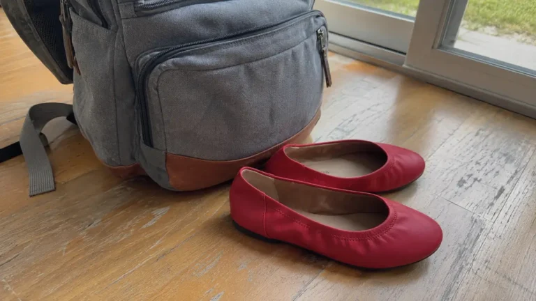 Bright poppy red Amazon Essentials Ballet Flats resting on a wooden floor next to a travel backpack for Stylue.
