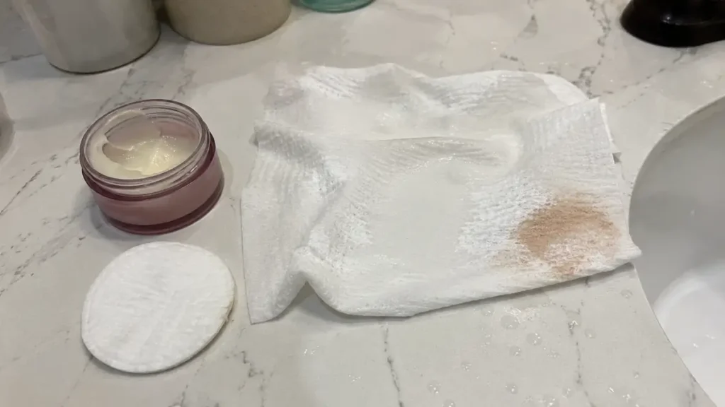 A facial towel, cleansing balm, and cotton pad arranged on a bathroom counter for makeup removal