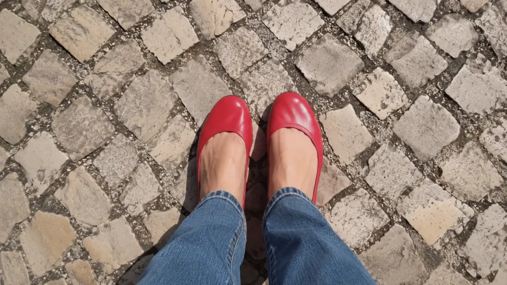 A person wearing bright red flat shoes standing on an uneven cobblestone street.